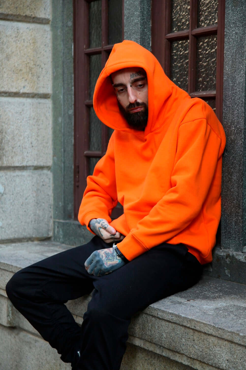 Plain multi colored hoodies featuring an orange design worn by a man sitting on a stoop.